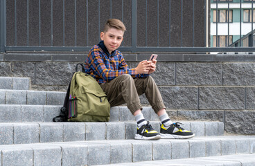 Cheerful boy schoolboy runs with a briefcase down the street. cute happy boy is reading using mobile phone. Back to school concept.