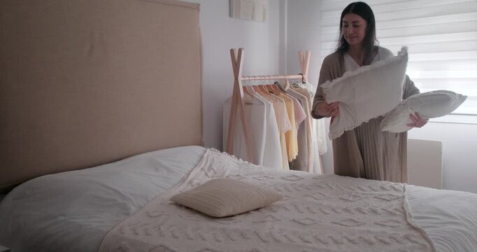 Cheerful dark haired woman arranging pillows and making bed in bright bedroom. Female doing housekeeping routine at home.