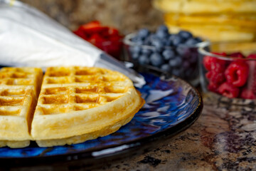 A golden waffle rests on a striking blue plate next to a piping bag and bowls of fresh berries, ready for final plating and serving in Waukesha County.