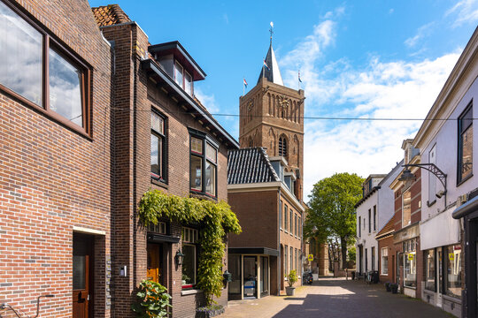 View of rustic brick buildings flank a narrow street leading to a towering church steeple under a vibrant blue sky, Noordwijk, South Holland, Netherlands.