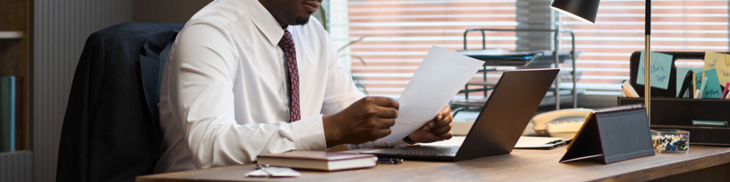 Middle aged Black man sitting at desk reviewing documents in office setting, wearing business attire, working with laptop and paperwork, embodying role of school principal