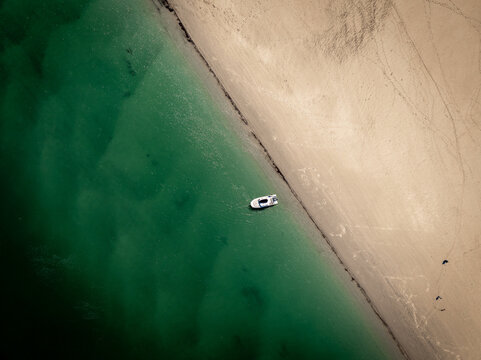 Aerial view of a lone boat resting on the edge where the dark teal water meets the tan sandy beach, Gloucester, Massachusetts, United States.
