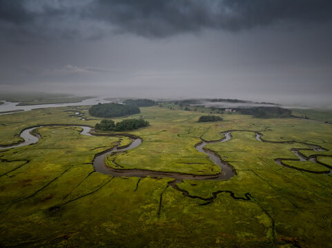 Aerial view of winding waterways carve through the vibrant green marshland under a brooding, grey sky, Gloucester, Massachusetts, United States.