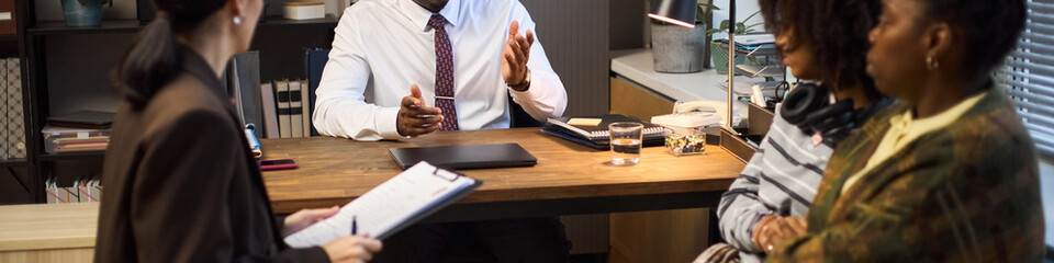 Middle aged Black man sitting at desk talking to two middle aged Black women and one middle aged Asian woman during school principal meeting, discussing documents and addressing concerns