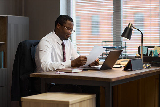 Middle aged Black man sitting at desk reading documents, working with laptop and tablet in modern office, appearing focused and professional, school principal performing administrative tasks