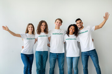 A group of smiling millennial volunteers stands together against a light wall, showing enthusiasm...