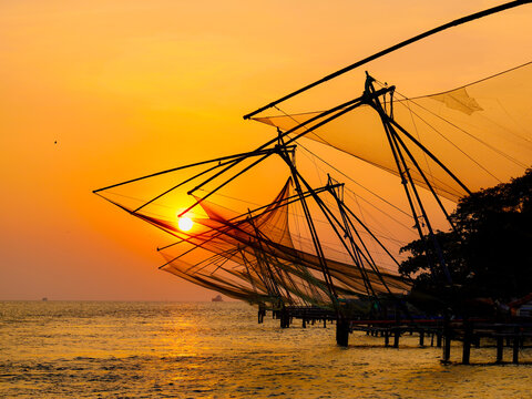 View of the intricate Chinese fishing nets silhouetted against the fiery sunset, casting a golden glow over the tranquil waters, Kochi, Kerala, India.