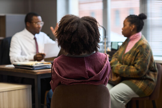 Black man appearing middle aged sitting at desk talking to Black woman and Black child in office setting, principal discussing school matters with parent and student