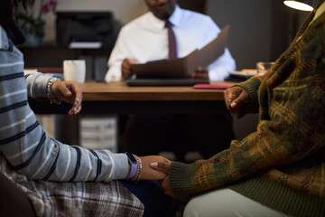 Two young adult women sitting close together holding hands in foreground, Black middle aged man sitting at desk in background reading documents, school principal context