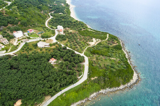 Aerial view of emerald trees meeting the turquoise sea, winding roads carving through lush landscapes, and buildings dotting the terrain, Paralia, Preveza, Greece.