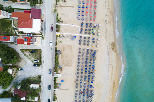 Aerial view of vibrant beach umbrellas punctuate the soft sand meeting the turquoise sea next to a road with cars and buildings, Paralia, Preveza, Greece.