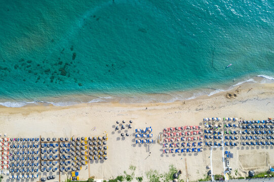 Aerial view of turquoise waters meeting the sandy beach lined with colorful umbrellas, creating a vibrant contrast of hues and textures, Paralia, Preveza, Greece.