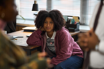 Black teenager girl sitting at desk in school principal office resting head on hand, looking at adult woman in foreground with thoughtful expression during conversation