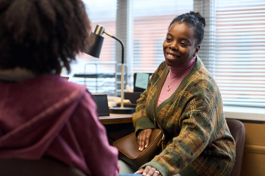 Middle aged Black woman principal smiling and talking to student in office, sitting at desk with hand resting on student arm, engaging in supportive conversation during school meeting