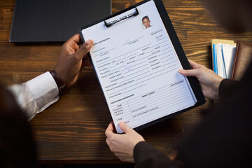 Caucasian woman holding clipboard with personal data sheet while Black man sitting across table reviewing document, both appearing as middle aged professionals in school principal office setting