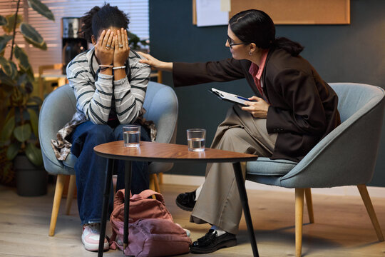 Middle aged Caucasian woman comforting upset Black teenager covering face with hands during counseling session in school office, both sitting in chairs with clipboard and glasses of water on table