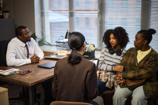 Middle aged Black man sitting at desk talking with middle aged Black woman, Black teenage girl and Black young adult woman during school principal meeting in office setting