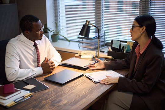 Middle aged Black man sitting at desk listening to middle aged Caucasian woman discussing documents in office setting, both engaged in professional conversation during school principal meeting