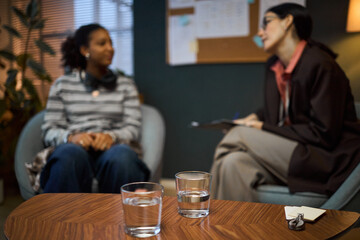 Middle aged Caucasian woman interviewing young Black woman in office setting, both seated and engaged in conversation, glasses of water and notepad on table in foreground
