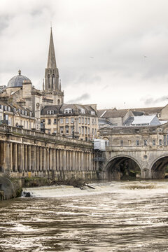 View of the Pulteney Bridge arches gracefully over the rushing Avon River, under a cloudy sky, with the city's honey-colored buildings, Bath, England, United Kingdom.