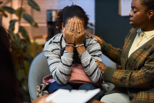 Young adult Black woman sitting with head in hands, being comforted by another young adult Black woman placing hand on shoulder in school principal office during emotional conversation