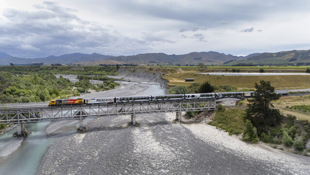 Aerial view of a train crossing a bridge over a wide, shallow riverbed, contrasting with the lush greenery and distant mountains, Seddon, Marlborough Region, New Zealand. - Powered by Adobe
