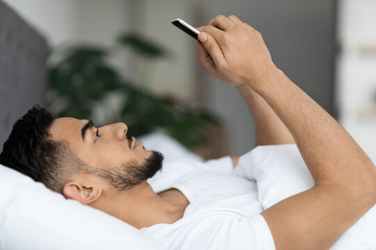 A young Arab man is lying in bed, using his smartphone to browse the internet. He appears relaxed in a cozy bedroom, showcasing modern decor. The focus is on his engaged expression.