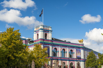 View of a vibrant building with bold pink, purple, and blue accents under a blue sky dotted with clouds, framed by green trees, Indre By, Denmark.