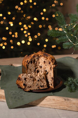 Traditional Christmas panettone sliced on plate, green linen napkin and pine tree branch on wooden table, blurred Christmas tree lights on background. Festive holiday baked fruitcake with raisins