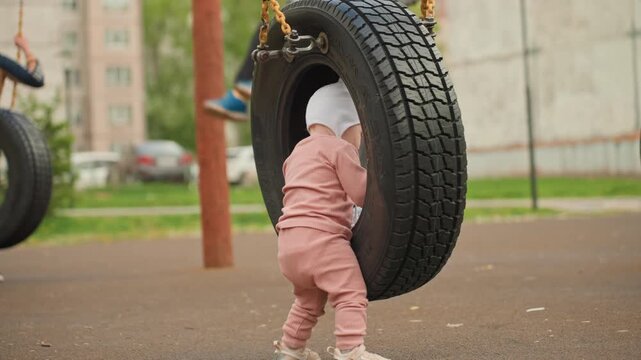 Pinkclad Youngster Cautiously Probes Swing, Toddler Dressed In Pink Examines Tire Swing With Playful Hesitation, Little Girl Wearing Pink Takes Tentative Steps To Explore Playground Swing Carefully