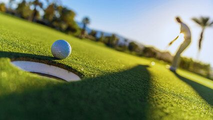 Golf ball on hole edge with golfer putting.