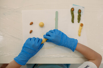 Top view of child's hands in blue gloves shaping dough log into "witch finger" cookie. Finished finger shapes topped with almonds as fingernails on table. Hands-on children's baking workshop.
