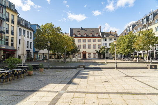 View of a large cobblestone Am Plan square bordered by colorful buildings and mature trees under a bright sky, Koblenz Mitte, Rhineland-Palatinate, Germany.
