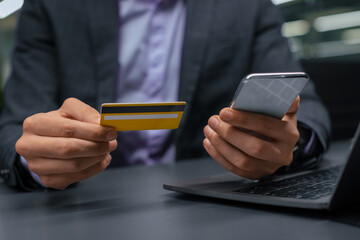 A person holds a credit card in one hand while using a smartphone in the other hand. The individual is seated at a desk with a laptop in front, engaged in online payment activity.
