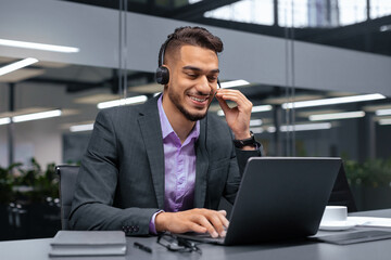 A man sits at a desk in a modern office, wearing a headset. He smiles while typing on his laptop. The workspace has large windows and indoor plants in the background.