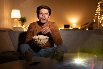A man sits on a sofa at home, shocked while watching a movie with a domestic projector in the...