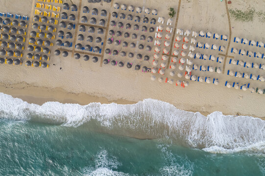 Aerial view of vibrant sunshades dotting the sandy beach where turquoise waves crash, creating a stunning contrast of colors, Vrachos, Preveza, Greece.
