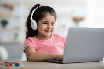 A happy young girl sits at a desk at home, wearing headphones and focused on her laptop. She is actively participating in an online learning session.