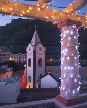 View of a church steeple rising above the rooftops, framed by a festive trellis adorned with twinkling lights, casting a warm glow in Ponta do Sol, Madeira, Portugal.