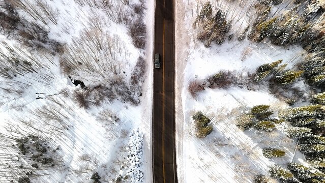 Aerial view of a lone car navigates a stark asphalt road dividing the snow-laden forest, the trees casting long shadows on the fresh snow, Mesa, Colorado, United States.