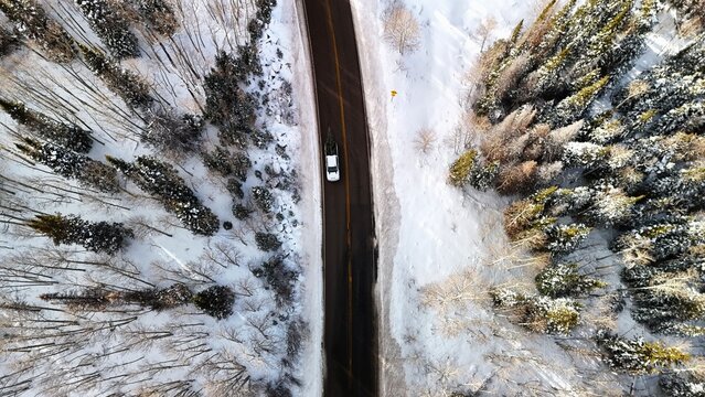 Aerial view of a solitary car winds its way through a snow-laden road, cutting a dark swathe through the pristine white landscape surrounded by snow-covered trees, Mesa, Colorado, United States.