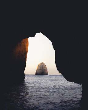 View of a solitary rock formation rising majestically from the tranquil ocean, framed by the dark, cavernous mouth of a coastal cave, Lagos, Faro District, Portugal.