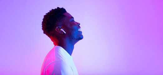A cheerful young black man listens to music with earphones, eyes closed in a vibrant neon light. He...