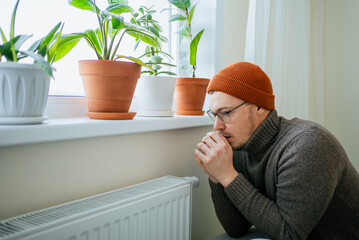 Man suffering from cold indoors due to high heating costs, keeping warm in a hat and sweater while trying to save money on expensive utilities, depicting energy crisis and home comfort issues