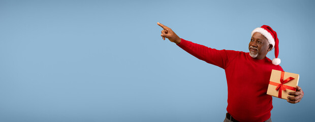 A man wearing a red sweater and a Santa hat stands against a blue backdrop. He holds a wrapped gift in one hand while pointing to the right with the other hand.