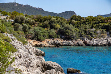 Scenic rocky bay with turquoise clear water revealing stony seabed, forested foothills under a bright sky. Idyllic Mediterranean coastal escape. Kaş, Turkey, Mediterranean.

