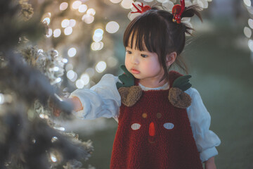 Cute asian little girl in reindeer costume and santa hairpin touching christmas tree with warm light bokeh for holiday season and childhood joy concept