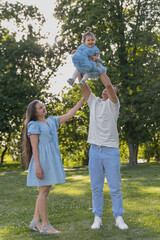 Fototapeta premium A young Caucasian man lifts a toddler girl in a blue dress. A young Caucasian woman stands beside them, smiling. They are in a green park with trees.