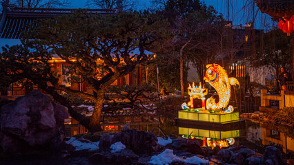 View of a glowing koi lantern casts a warm reflection on the dark water of a serene pond in a Chinese garden at night, Portland, Oregon, United States.