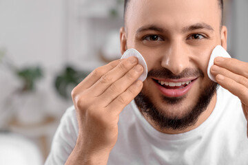 Handsome young happy man cleansing his face with micellar water and cotton pads in bathroom
